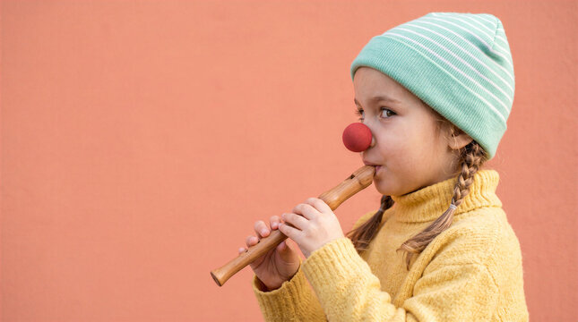 A young girl with braids wearing a striped beanie and a red clown nose playing a wooden recorder against a solid orange background