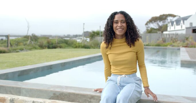 Adult African woman sitting on pool coping in mustard top, camera moving closer, turning, smiling