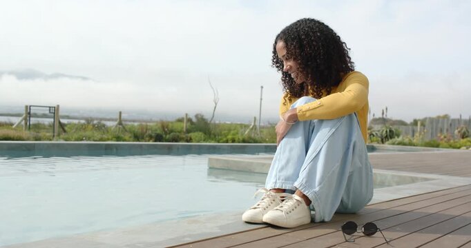 Woman watching water, sitting poolside on wooden deck, touching, splashing water, sunglasses nearby