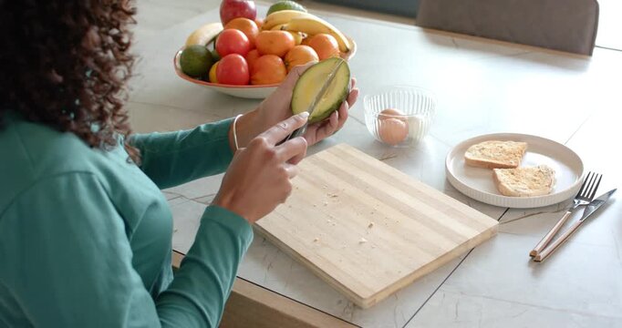 African American woman in teal steadying halved avocado and scraping with knife preparing toast
