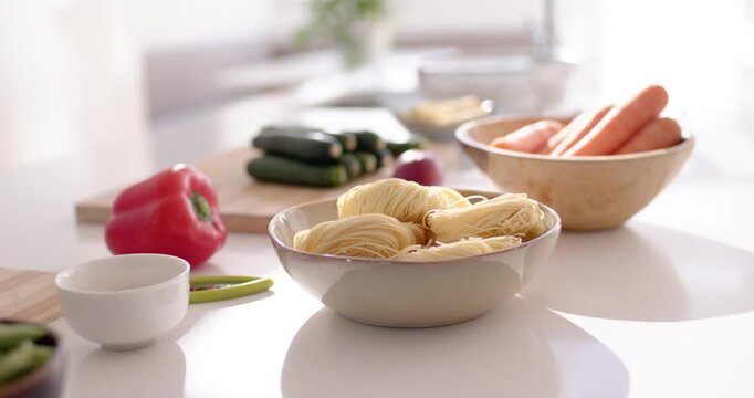 Camera is pushing and pulling, shifting focus around pale bowl of dry noodles, revealing meal prep
