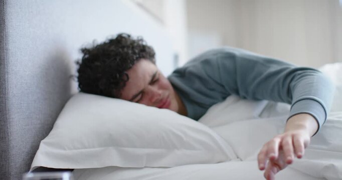Adult male in bed reaching for ringing white alarm on nightstand with water glass, pulling duvet