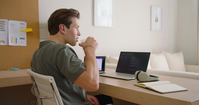 Man at desk green tee seeing tablet, reaching, lifting mug, stretching to relax, resuming laptop