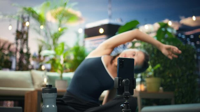Woman adjusts a tripod-mounted smartphone from a rooftop yoga mat, preparing to film an online fitness and yoga tutorial with mobile recording equipment.