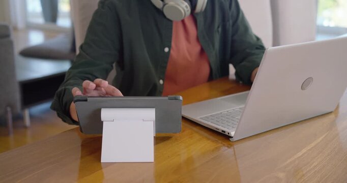 After opening laptop, remote worker is typing and tapping tablet on stand at wooden table