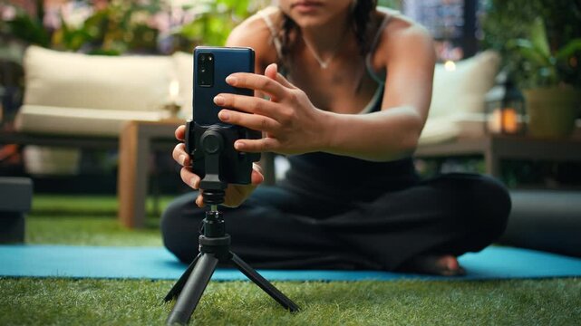 Woman leans forward to position a smartphone on a tripod beside a rooftop yoga mat, preparing for a self-recorded outdoor fitness demonstration and adjusting her mobile video setup.