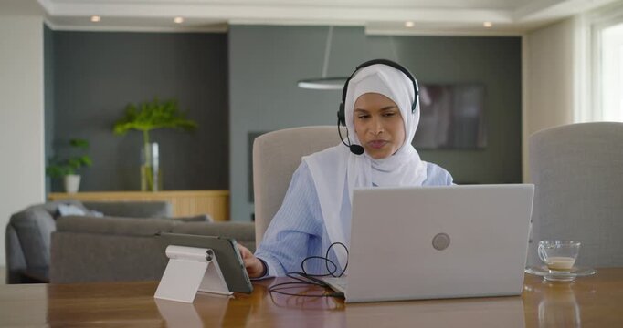 Woman in hijab joining remote meeting at home, adjusting tablet and headset, typing laptop