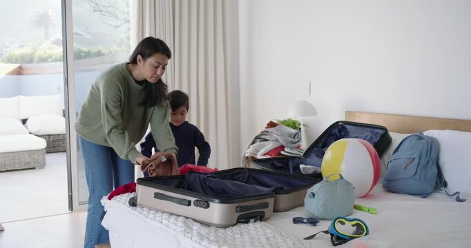 Asian mother and son packing on bed preparing for trip, folding clothes into suitcase