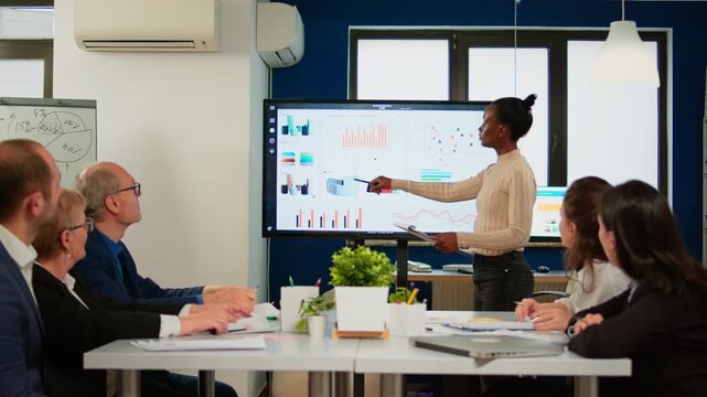 Analytics, presentation and businesswoman explaining charts on a digital display to seated colleagues, with team recognition and applause during a business briefing on performance and strategy.