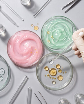 Cosmetic science and laboratory research concept, gloved hand using pipette over petri dishes with colorful pink, green and oil gel textures on grey background.