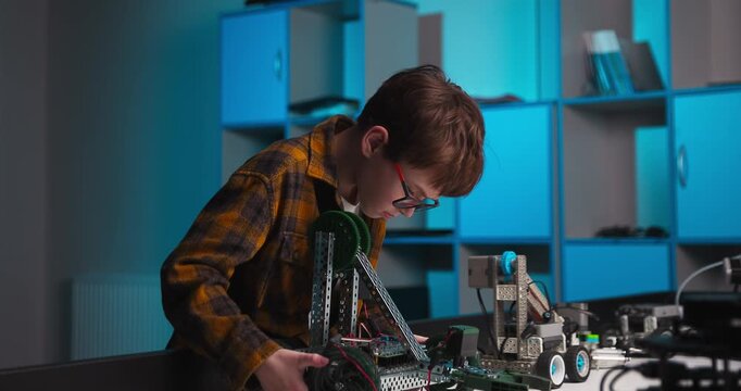 Young student assembling and adjusting a wheeled robot beside another robotic vehicle during a hands on engineering project in a modern workshop