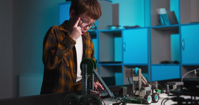 Young student adjusting a wheeled robotic vehicle prototype on a workshop table during hands on robotics project in a modern technology classroom setting
