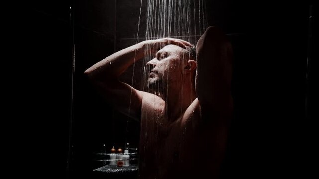 Dark shower. Rain falls. Man rinses his head. He closes his eyes. Water flows. Body glistens. Emotionless. Calm. Quiet. Use for spa, wellness, or relaxation themes