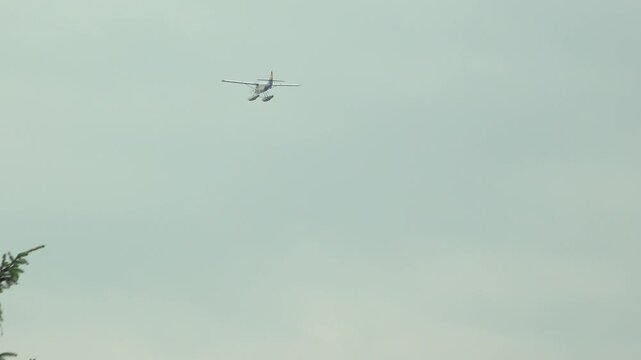 A propeller floatplane soaring high through the air in Vancouver, British Columbia. The small commercial aircraft flies over a coastal bay under an overcast summer sky.