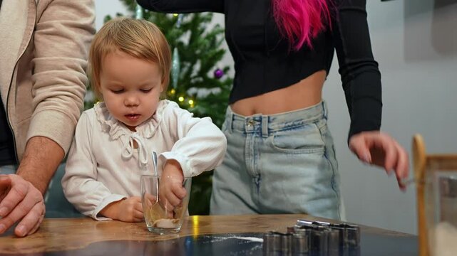Little girl is sitting at the table and pouring finger into cinnamon then licking it to try the taste while her family is cooking pastries together. Small child engaged in process of cooking biscuits