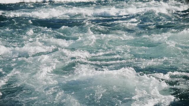 Churning water surges with force and energy. White foam crashes against deeper turquoise waves. Motion suggests powerful currents or a vessel's wake