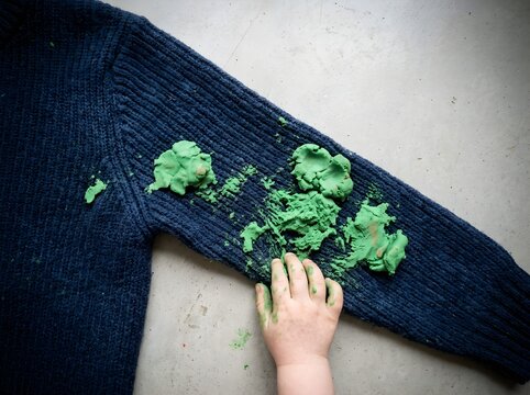 Child hand pressing bright green play dough directly onto dark blue knitted sweater sleeve. Visual metaphor for inevitable domestic mess stress and parenting chaotic reality.