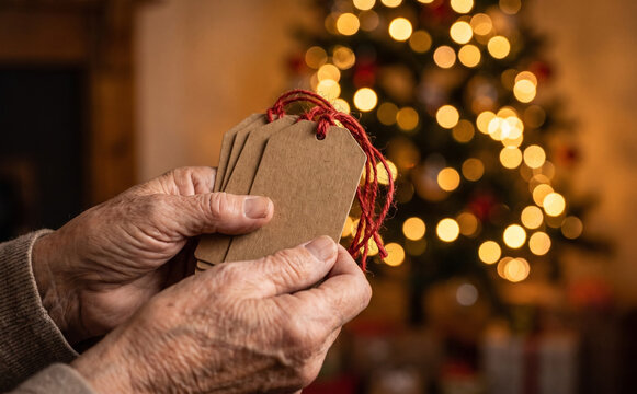 Senior adult hands holding blank brown paper gift tags with red string in front of a Christmas tree
