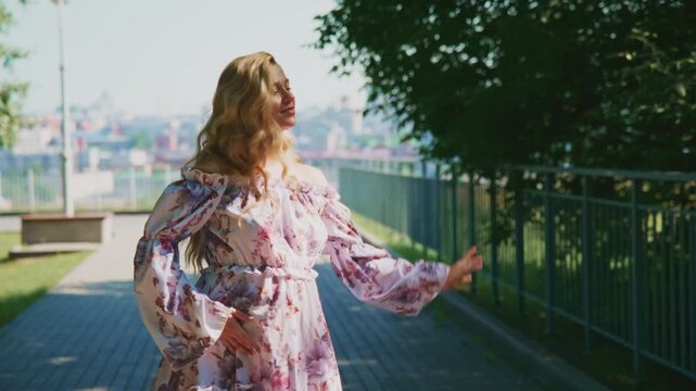 Young woman walks barefoot on sunny sidewalk. Her floral dress flutters gently with breeze. She smiles softly, enjoying city views beyond trees. Natural light highlights her long, wavy hair