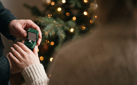 Man proposing to woman with engagement ring in green velvet box in front of glowing Christmas tree