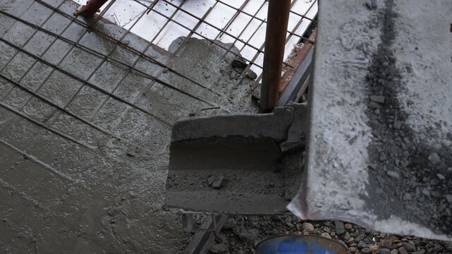 Wet concrete pours from a chute onto a reinforced steel grid, slowly spreading to fill the space below a blue barrel and scattered gravel in a stationary construction site shot.
