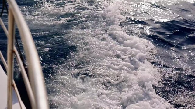 White foam trails behind a moving boat. Sunlight glints off deep blue ocean waves. Railing edge frames the sea's motion. Joyful journey across open waters. Perfect for travel, adventure