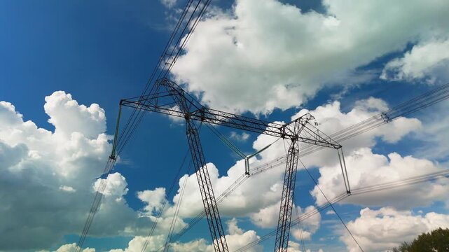 Steel towers stretch upward against a bright blue sky. Cables run taut, connecting distant power sources. Clouds drift slowly, casting soft shadows on metal structures. No people appear in this scene