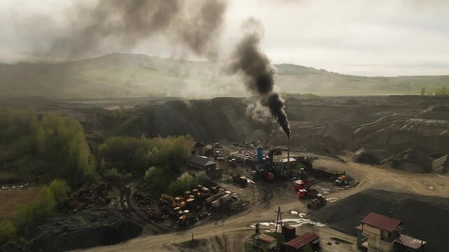 Black smoke billows from a mining site. Heavy machinery dominates the rugged landscape. Small structures dot the industrial zone. Workers move amidst the machinery