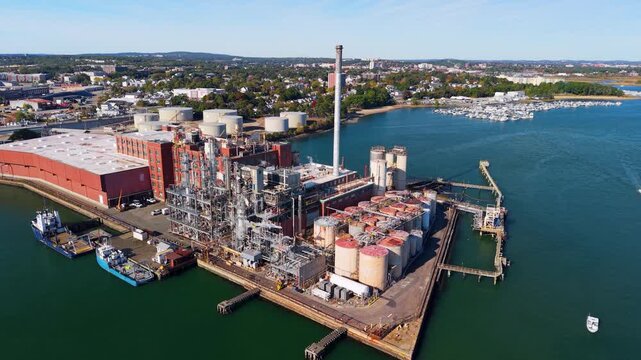 Aerial view of an industrial facility with storage tanks and a tall smokestack on the waterfront near the Fore River Bridge in Quincy, Massachusetts, United States.