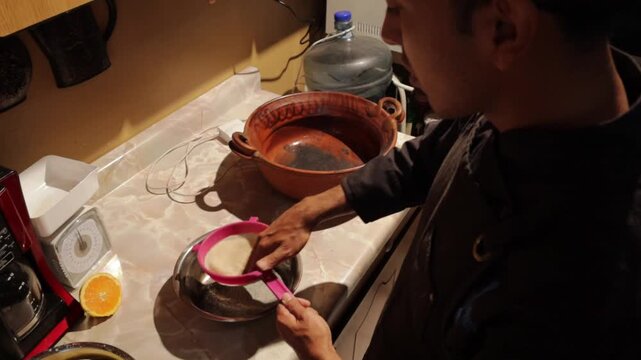 Hispanic man preparing traditional mexican amaranth dessert by straining ingredients through a pink sieve in a rustic kitchen at night with warm, intimate lighting