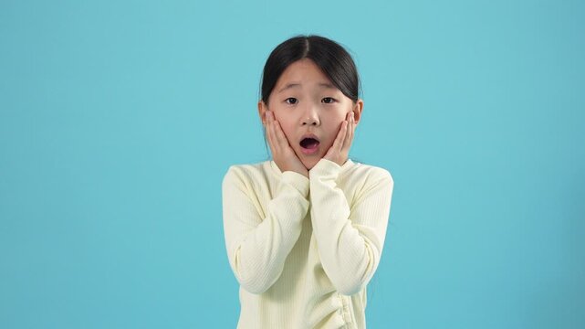 Shocked Young Asian Girl Holding Face in Disbelief Against Blue Background