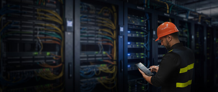 A technician wearing safety helmet using tablet beside server racks, checking network equipment and IT infrastructure during data center inspection and maintenance.