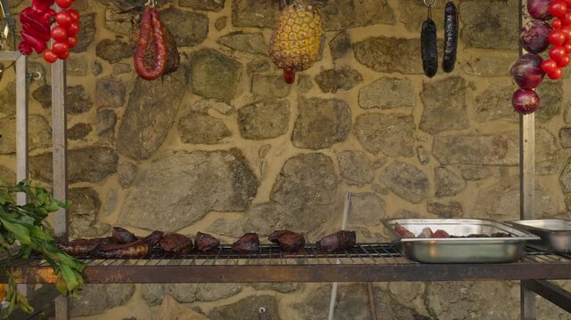 Outdoor grill station against a granite wall, with chorizo, blood sausage, pineapple, red onions, peppers and tomatoes hung above the cooked meat and trays