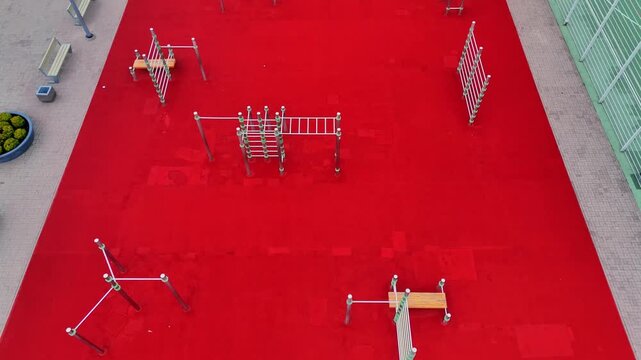 Aerial drone view of an empty calisthenics plaza with bodyweight stations, climbing frames and bars arranged across vivid red rubber flooring.