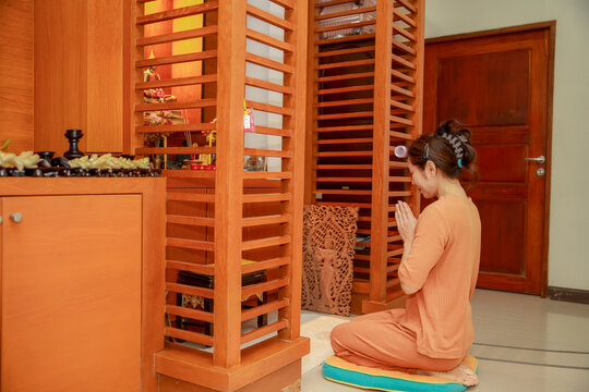 Asian woman praying with hands folded in front of wooden home shrine