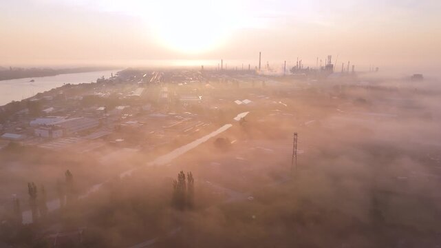 An aerial drone view of a sprawling industrial refinery and river bathed in the golden light of sunrise through morning fog. Perfect for energy sector and environmental themes.