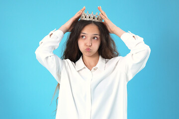 Expressive young woman adjusting gold tiara crown on head against blue background © Midea