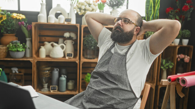 Man florist in apron leans back with hands behind head by laptop beside rolls of wrapping paper and flower shelves in studio; contemplation.