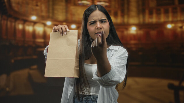 Young hispanic woman holds paper bag and points finger accusingly toward camera in studio, frowning and gesturing while demanding explanation; frustration accusation.