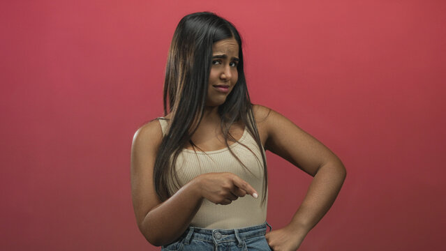 Young hispanic woman pointing finger to the left in a red studio, wearing beige tank top and jeans; skepticism.