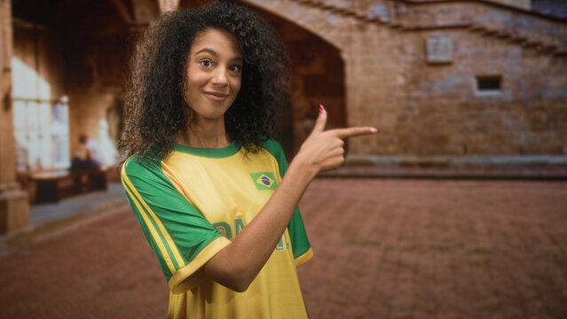 Woman with curly hair wearing brazil football team t shirt points finger with hand toward right in building courtyard while smiling; pride support celebration.
