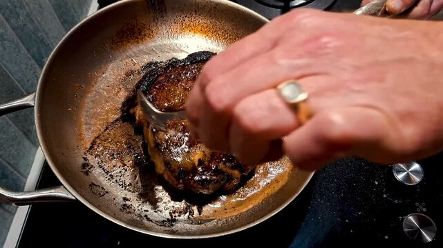 A man frying a big fat beef steak in a stainless steel pan on a gas hob and drenching it in butter with a spoon