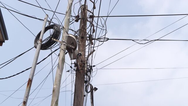 Utility poles carry tangled overhead wires, coiled cables and a satellite dish against a lightly clouded sky in Ghana.