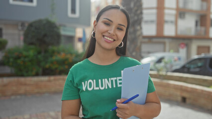 Woman holding clipboard and pen with visible hand, smiling in volunteer tshirt on a street;...