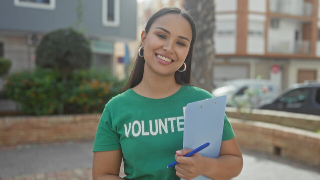 Woman holding clipboard and pen with visible hand, smiling in volunteer tshirt on a street; friendly community service.
