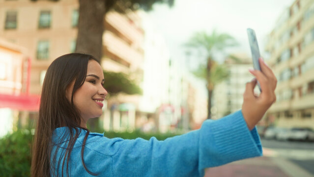 Young latin woman holding smartphone and smiling while taking a selfie on a city street; digital connection contentment.
