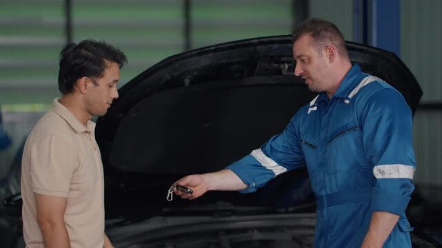 Two men stand by an open car hood. Mechanic in blue gestures to the engine. The customer shakes hands with the technician