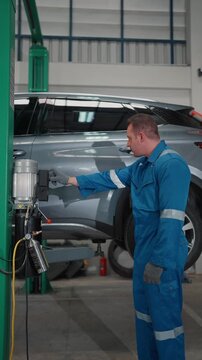 Technician wears blue coveralls in garage. He repairs a silver SUV's side panel. Equipment and tools surround him. He concentrates