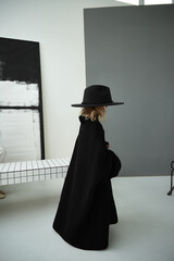 Full length profile portrait of a little girl wearing a dramatic oversized black knitted sweater and a wide-brim hat in an art studio. © Irina