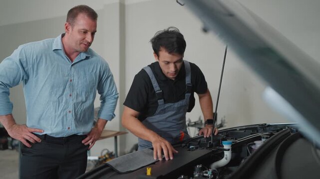 Two men in a garage, one in overalls, the other in casual wear. Mechanic gestures toward engine components while speaking. Client listens attentively